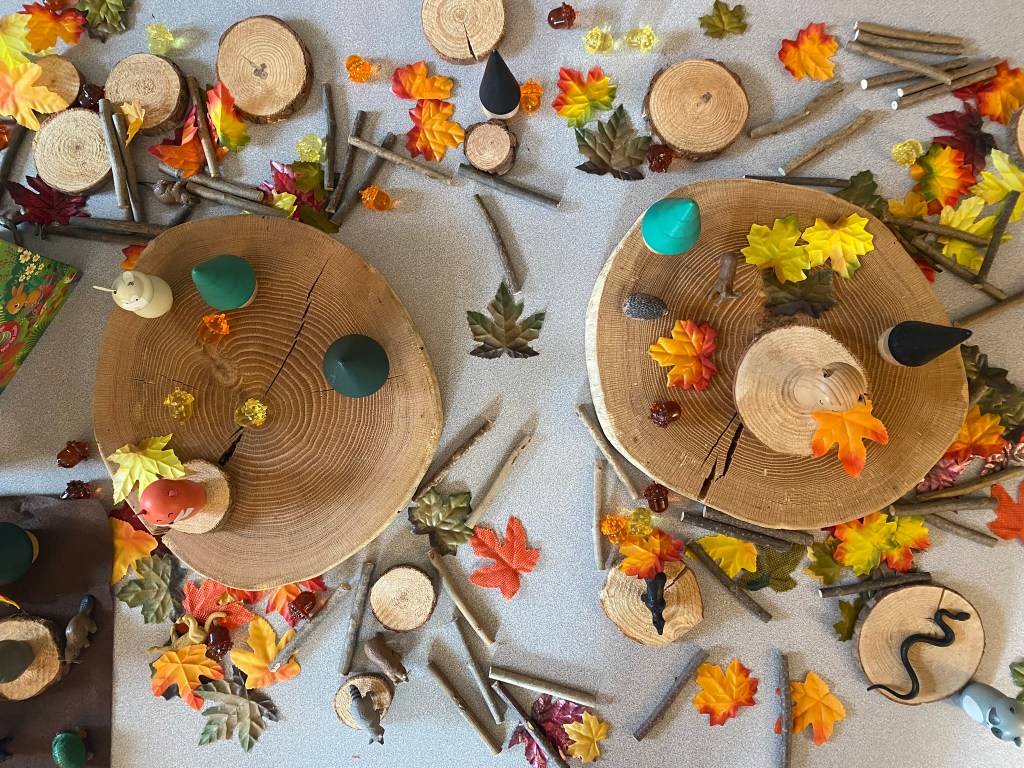 A tabletop arrangement featuring wooden rounds, colorful leaves, and decorative elements such as small animals and glass gems, creating a playful and natural scene.