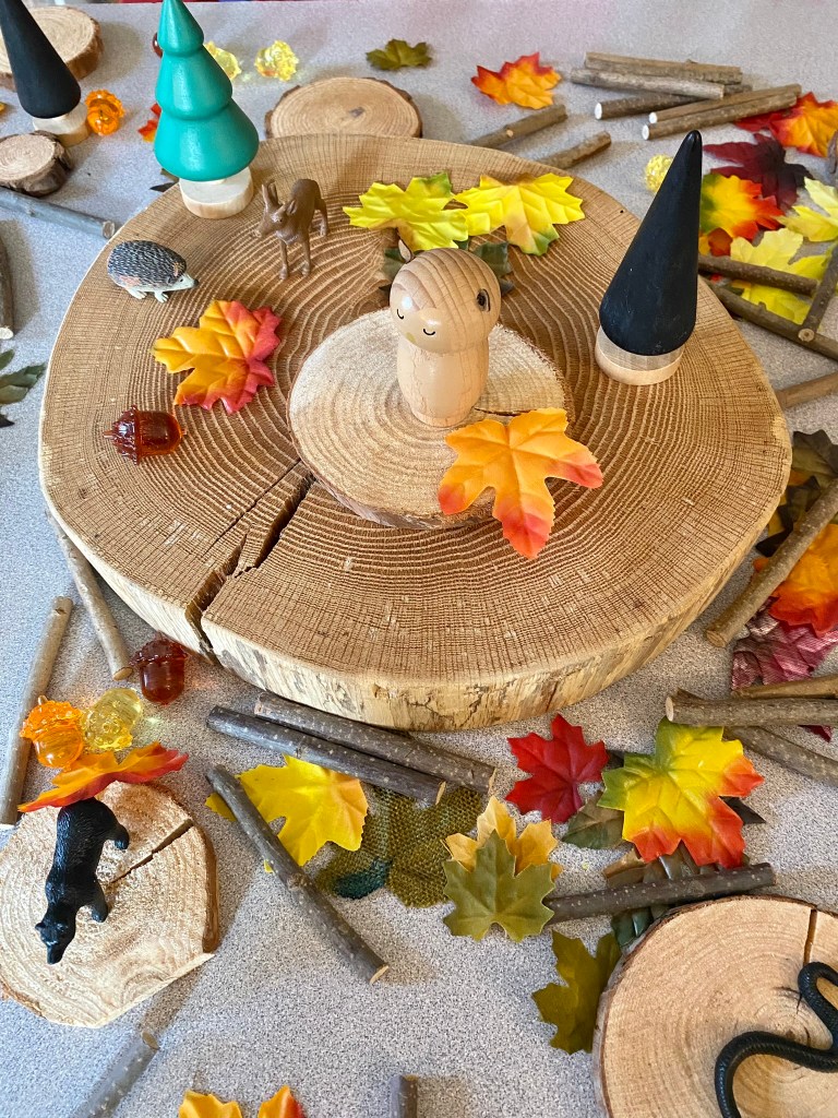 A wooden play scene featuring a round log base, colorful autumn leaves, small figurines including a hedgehog, deer, and a smiling wooden character, alongside decorative trees and twigs.