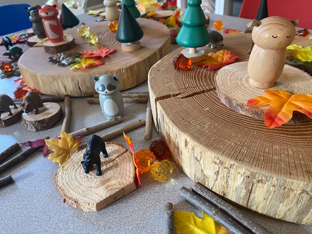 A colorful play scene with wooden animal figurines, trees, and autumn leaves arranged on large wooden slices and a tabletop.