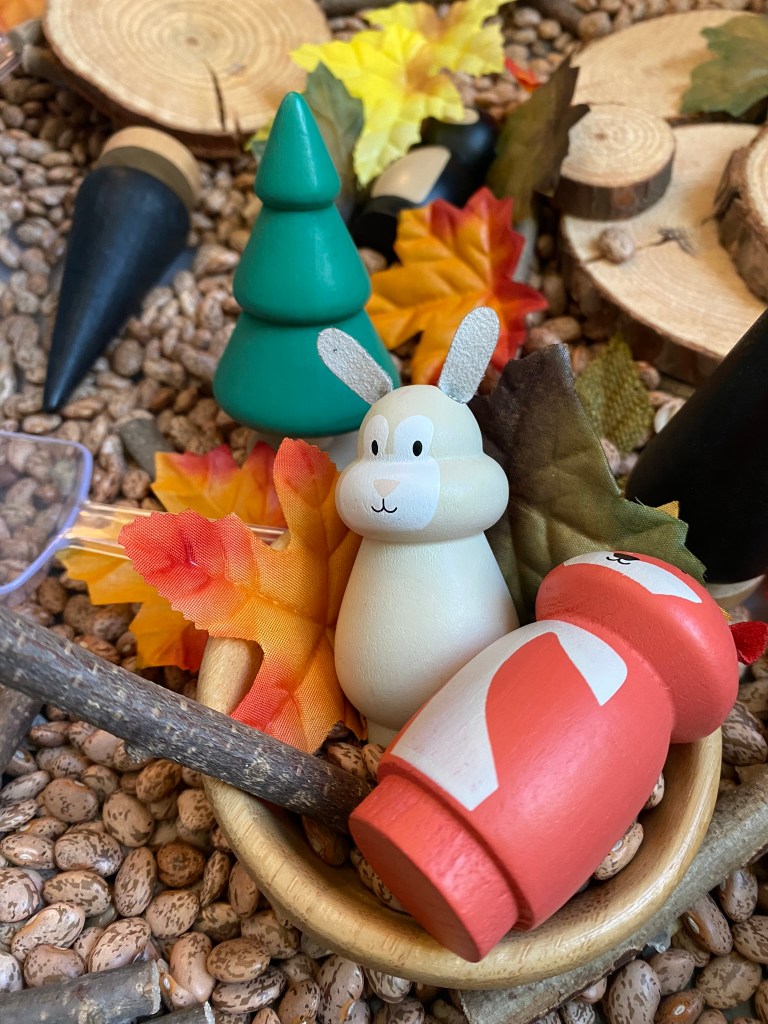 A bowl filled with colorful wooden toys, including a green tree, a white bunny, and a red mushroom, surrounded by artificial autumn leaves and pebbles.