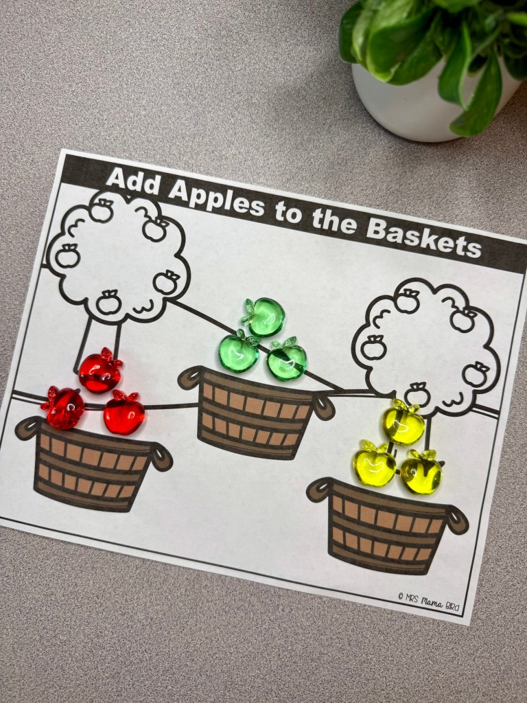 A colorful educational activity sheet titled 'Add Apples to the Baskets' featuring three illustrated baskets and apple counters in red, green, and yellow.