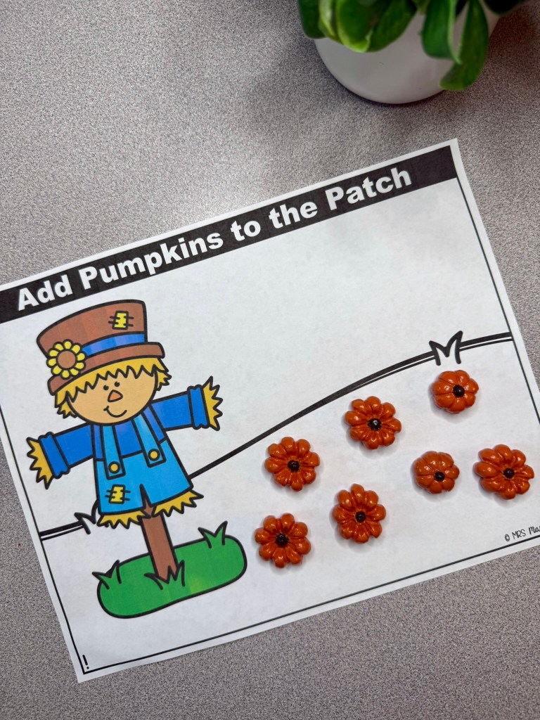 Printable worksheet featuring a cartoon scarecrow, with the text 'Add Pumpkins to the Patch' at the top and decorative orange flower shapes scattered below.