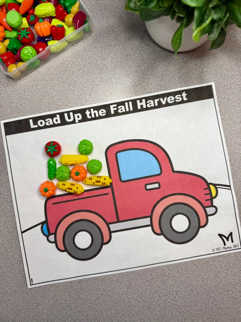 A colorful illustration of a red truck with a blue window, labeled 'Load Up the Fall Harvest,' filled with various plastic fruit and vegetable toys, placed on a gray tabletop.