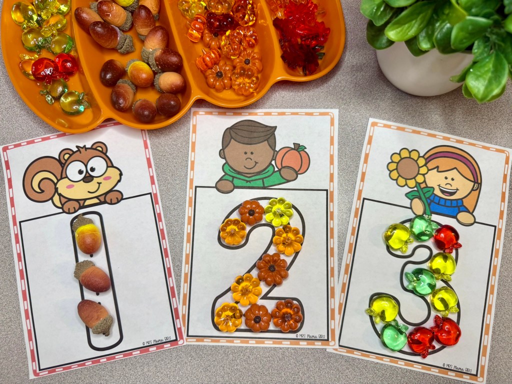 Three colorful counting cards with the numbers 1, 2, and 3, featuring playful illustrations of a squirrel, a boy with a pumpkin, and a girl with a flower. Each card is decorated with assorted counting manipulatives like acorns and colorful gems.